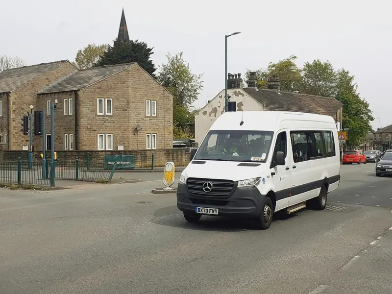 A white passenger minibus on a city street running a fixed TransportPlus shuttle route